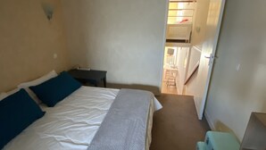 Een slaapkamer met een bed, nachtkastje, bureau en stoel. Er is een deur die leidt naar een keuken.