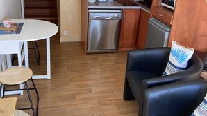 Een compacte keuken met een RVS vaatwasser, een magnetron en een koffiezetapparaat op het aanrecht. Er is een kleine eettafel met een stoel en een zwarte leren bank met een gestreept kussen.