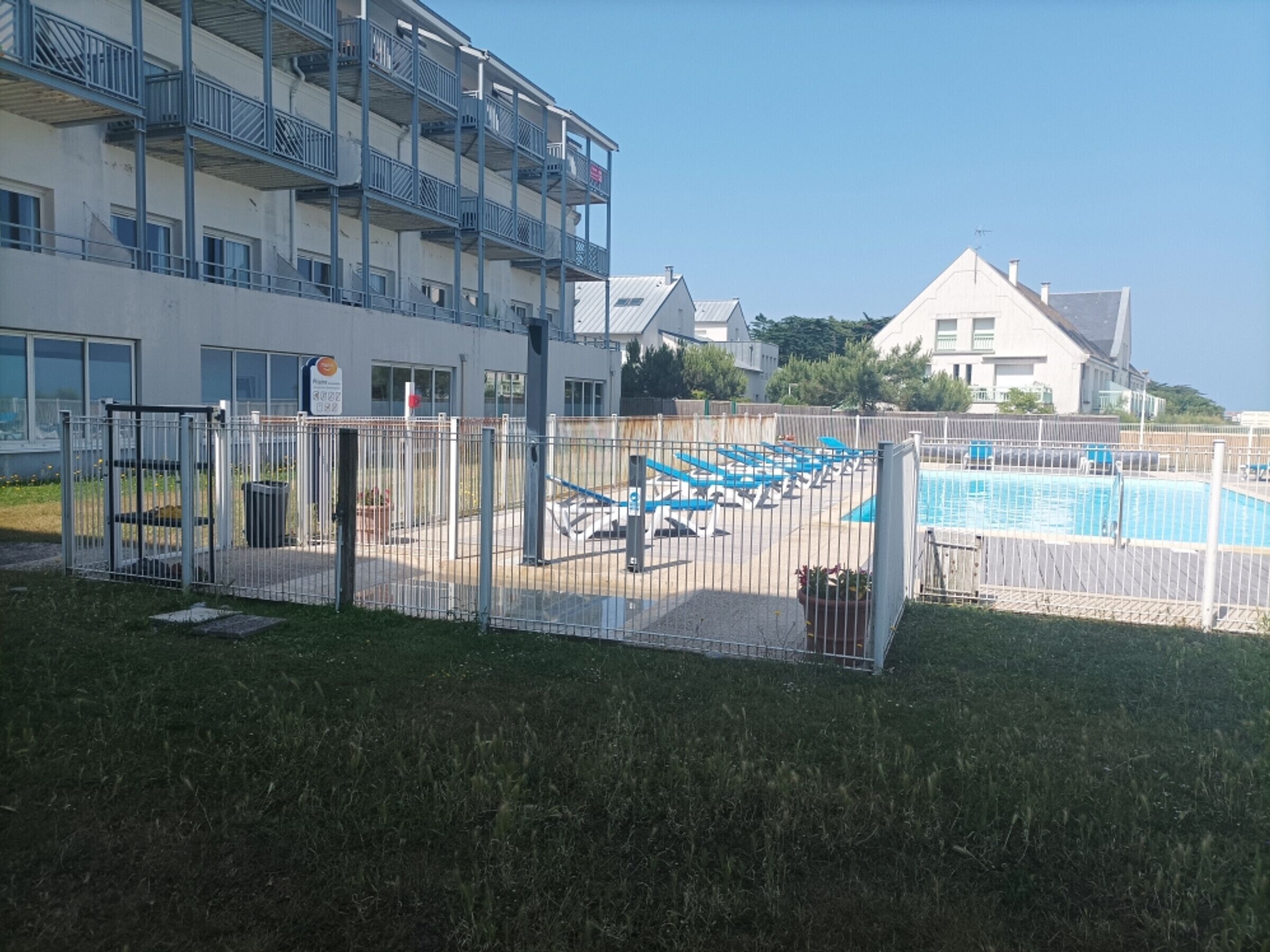 A fenced outdoor swimming pool area with lounge chairs and a building with balconies in the background.