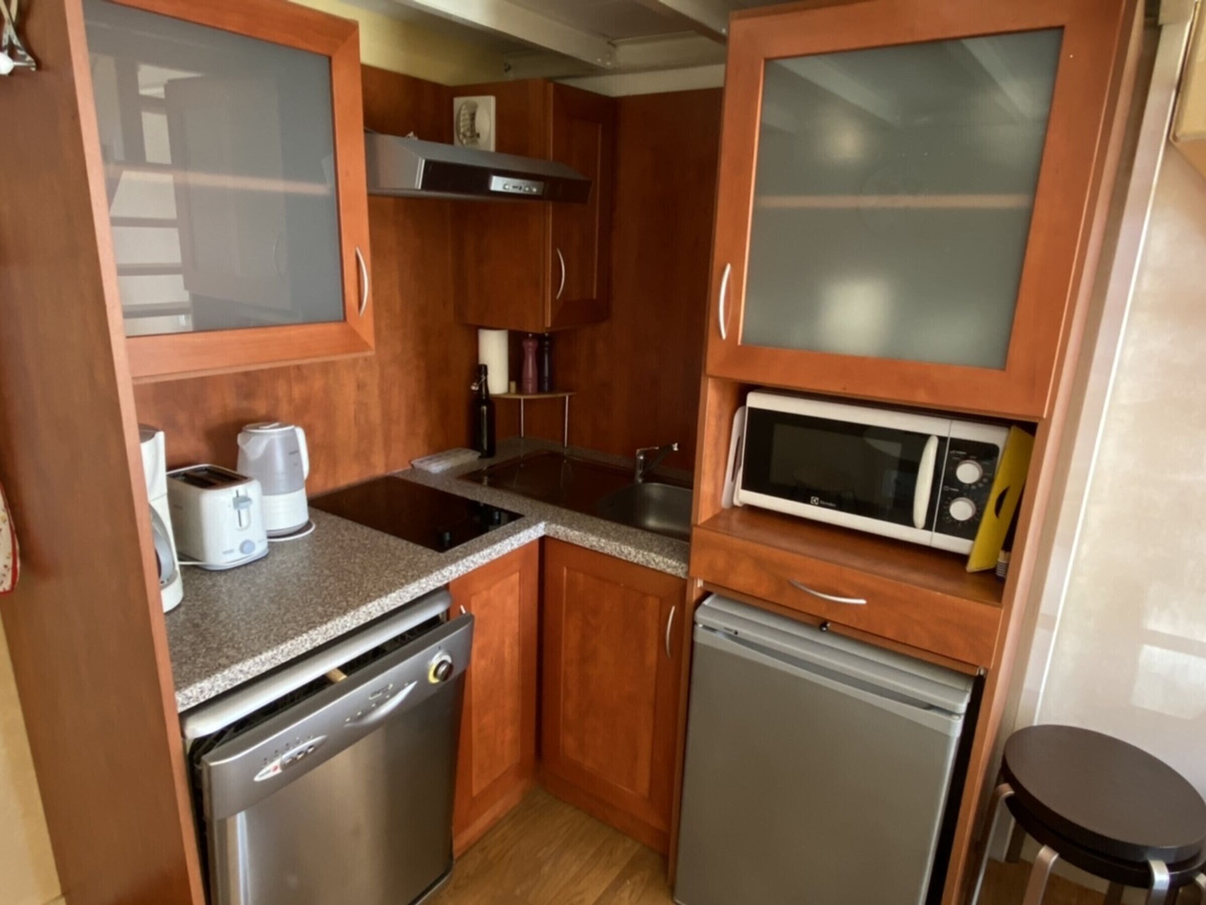 Una cocina compacta con armarios de madera, microondas integrado, lavavajillas y cafetera.