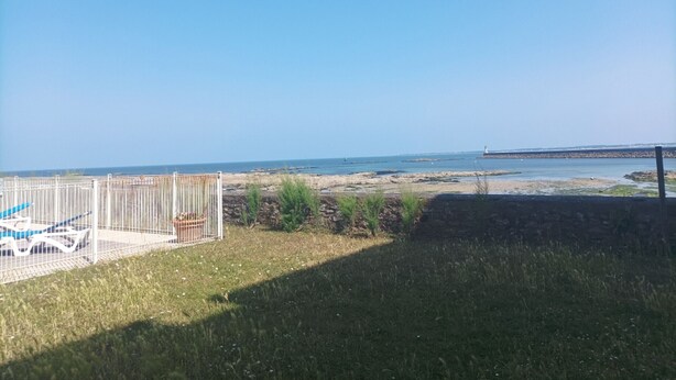 Een gazon met een witte hek en een metalen poort, leidend naar een strand met een rotsachtige kustlijn en een verafgelegen vuurtoren.