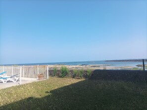 Ein grünes Feld mit einem weißen Zaun und einem Metalltor, das zu einem Strand mit felsigem Ufer und einem fernen Leuchtturm führt.