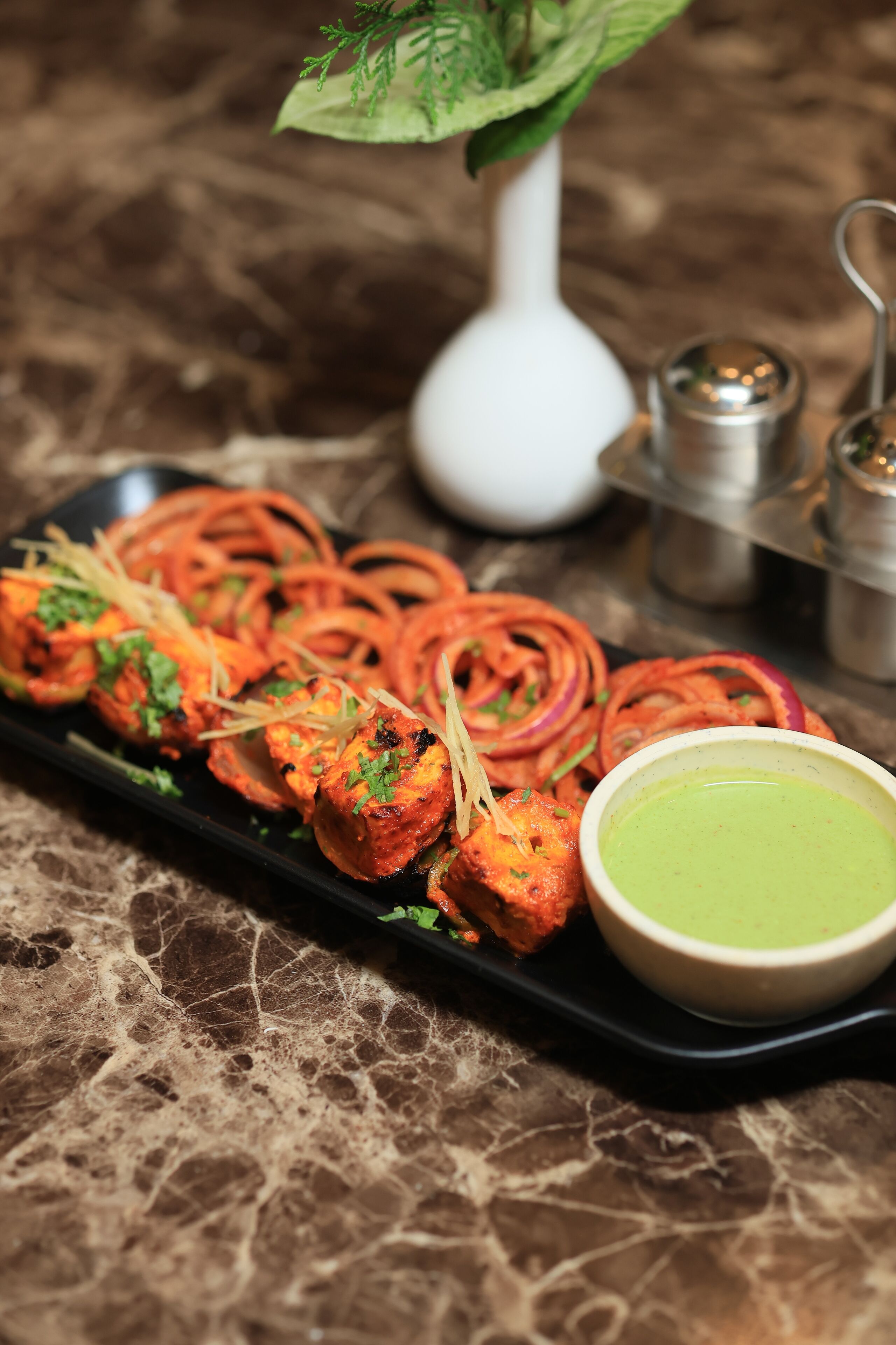 A black tray with tandoori chicken skewers, onion rings, and a bowl of green chutney on a marble surface.