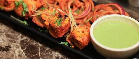 A black tray with tandoori chicken skewers, onion rings, and a bowl of green chutney on a marble surface.
