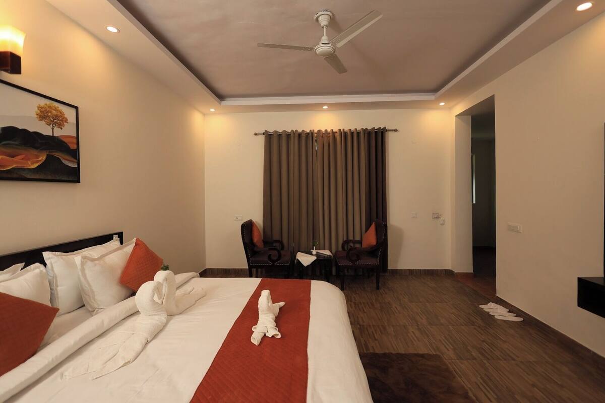 A hotel room with a bed, a desk, two chairs, a ceiling fan, and a painting on the wall.