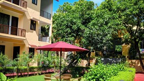A courtyard with a red umbrella, a round table, and chairs. There is a building with balconies and a tree in the background. - Chiraiya Resort and Hotels (Ramnagar)