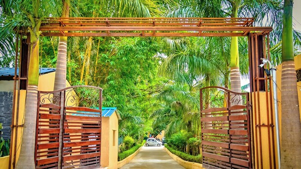 A gated entrance with a wooden archway, flanked by palm trees and a paved path leading to a parked car. - Chiraiya Resort and Hotels (Ramnagar)