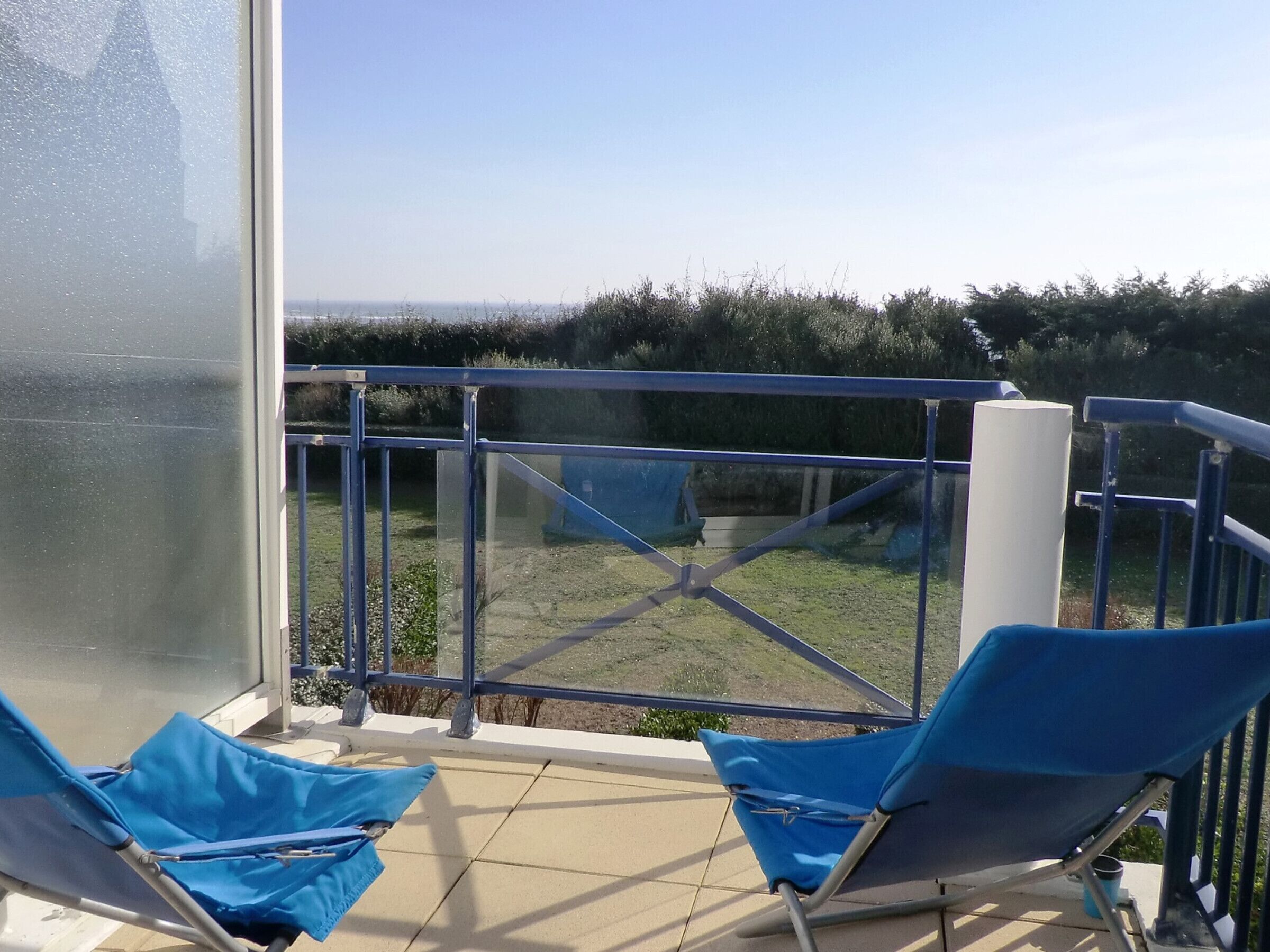 A balcony with two blue chairs, a glass door, and a view of a grassy area.