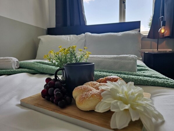 A neatly made bed with a wooden headboard, a bedside table with a lamp, a tray with grapes, a mug, and pastries on a green towel.