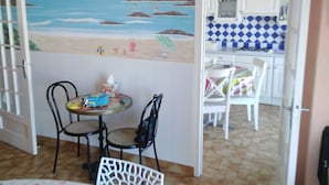Ein Essbereich mit Tisch und Stühlen, ein Gemälde mit Strandmotiv und eine Küche mit blau-weißen karierten Fliesen.