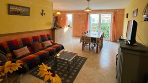 Una sala de estar con sofá, una mesa de centro, una mesa de comedor con sillas, un televisor sobre un mueble bajo, y una zona de cocina con mostrador y lavabo.