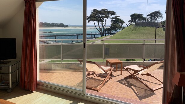 Une pièce dotée d’une grande porte-fenêtre coulissante qui donne sur une terrasse extérieure offrant une vue sur la mer et une colline verdoyante.