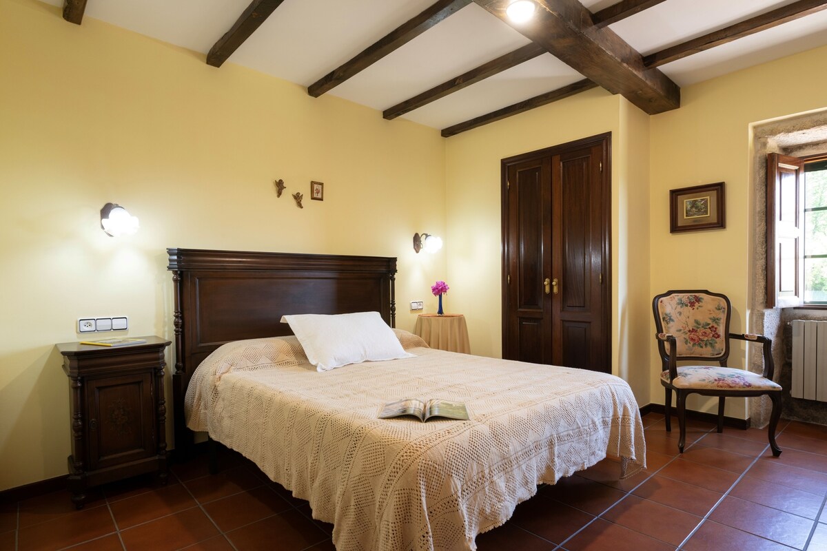 A bedroom with a large bed, wooden headboard, a nightstand, a chair, and a window with a view of greenery.