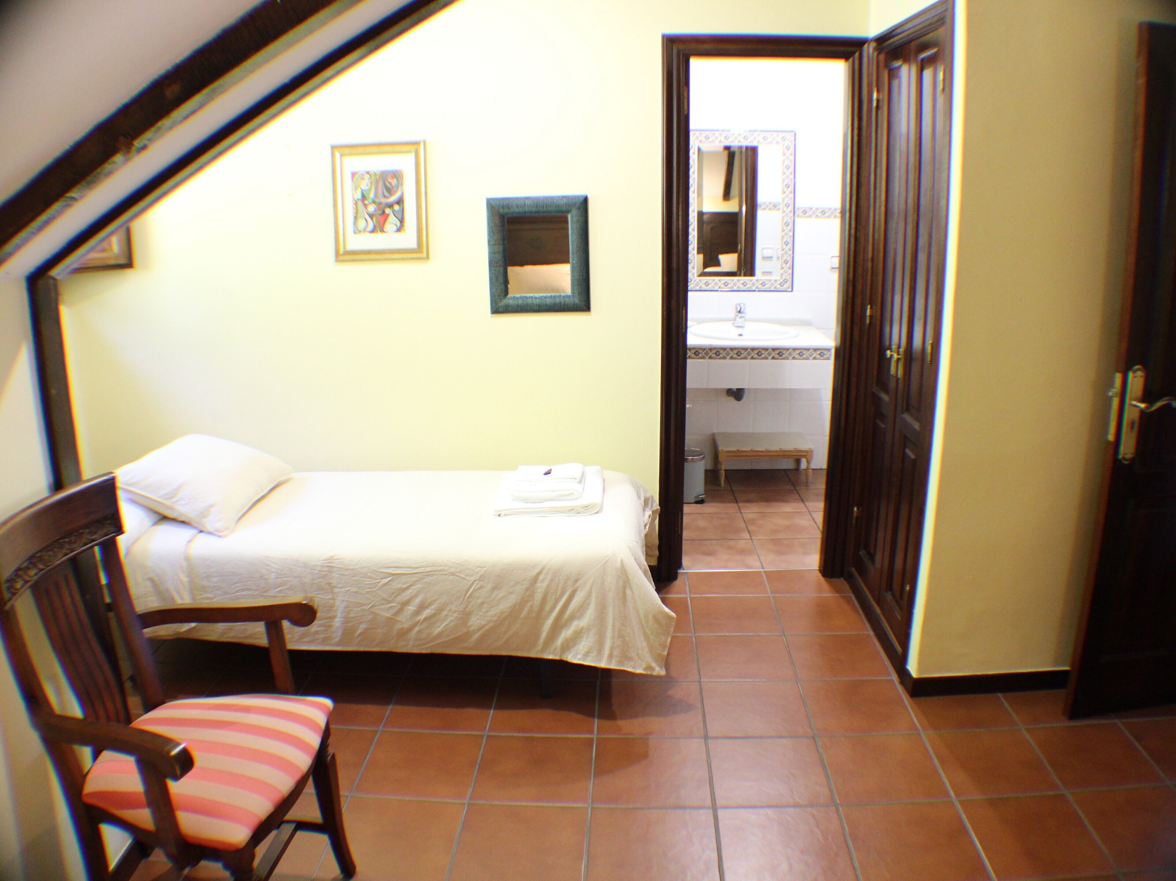 Un dormitorio pequeño con una cama individual, una silla, un cuadro enmarcado y un baño visible a través de una puerta abierta.