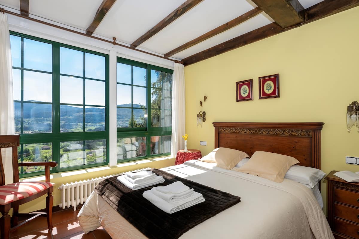 A bedroom with a large window, a bed with white linens, a wooden headboard, and a view of the countryside.