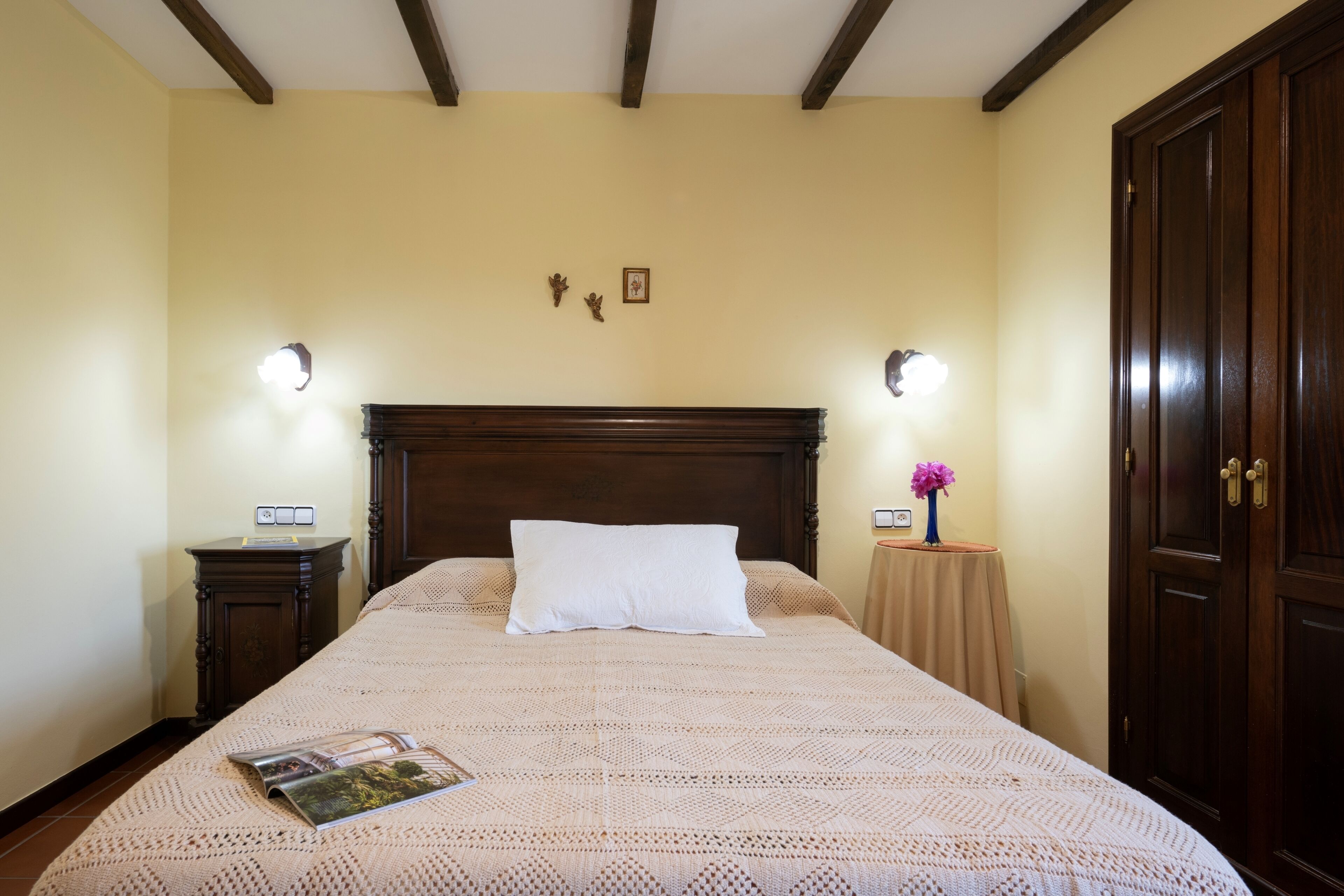 Een net opgemaakt bed met een wit kussen en een tijdschrift erop, een houten nachtkastje met een lamp, een kleine tafel met een bloevas, en een houten garderobe.