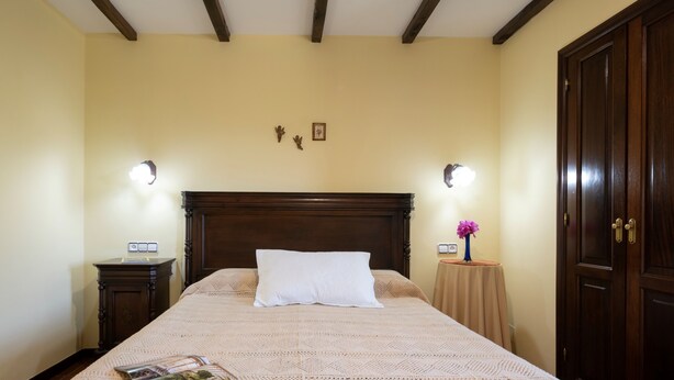 Un lit bien fait, avec un oreiller blanc et un magazine posé dessus, une table de chevet en bois avec une lampe, une petite table sur laquelle se trouve un vase de fleurs, et une armoire en bois.
