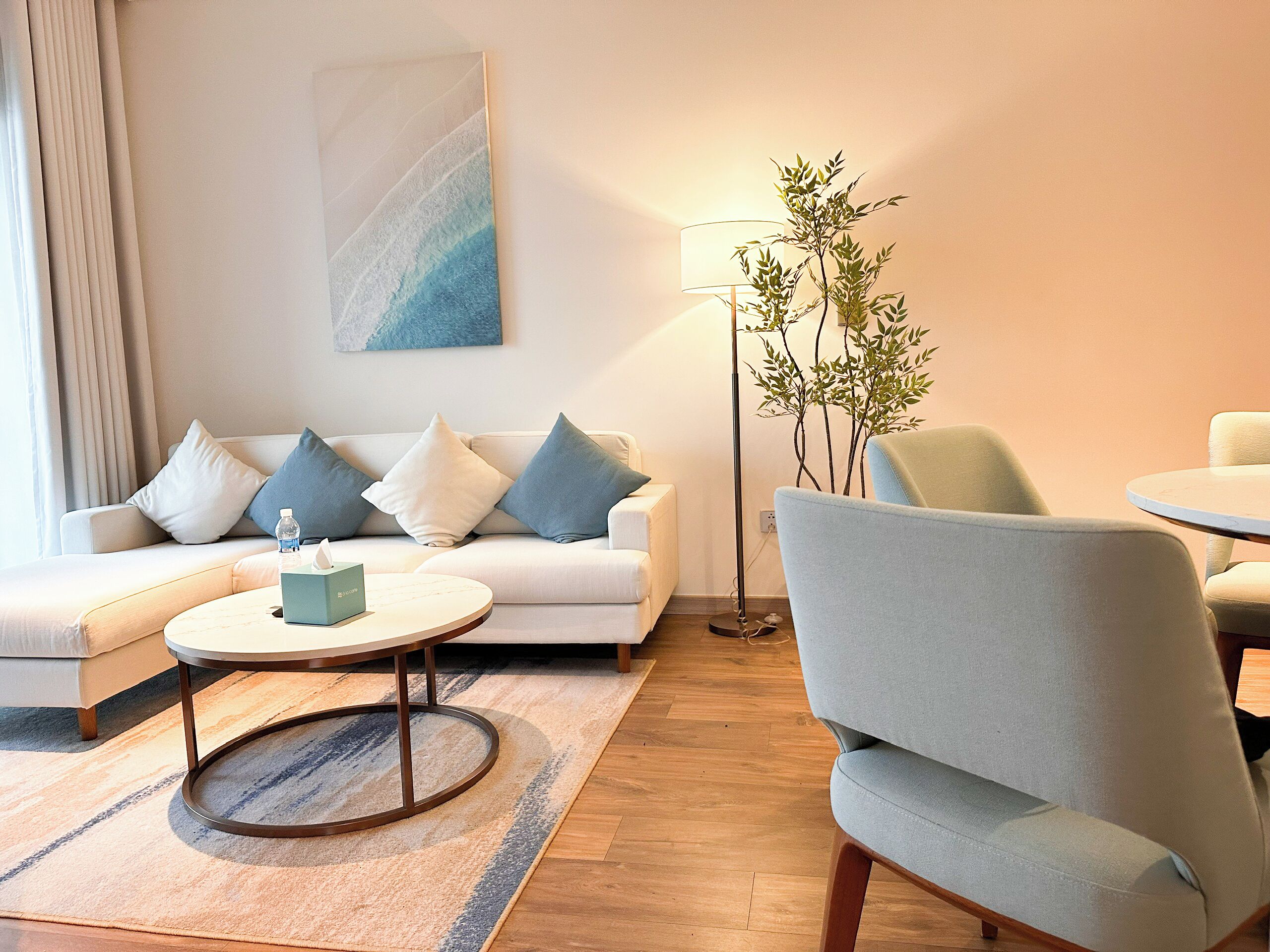 Ein modernes Wohnzimmer mit einem weißen Sofa, blauen und weißen Kissen, einem runden Couchtisch, einer Stehlampe und einer Topfpflanze.