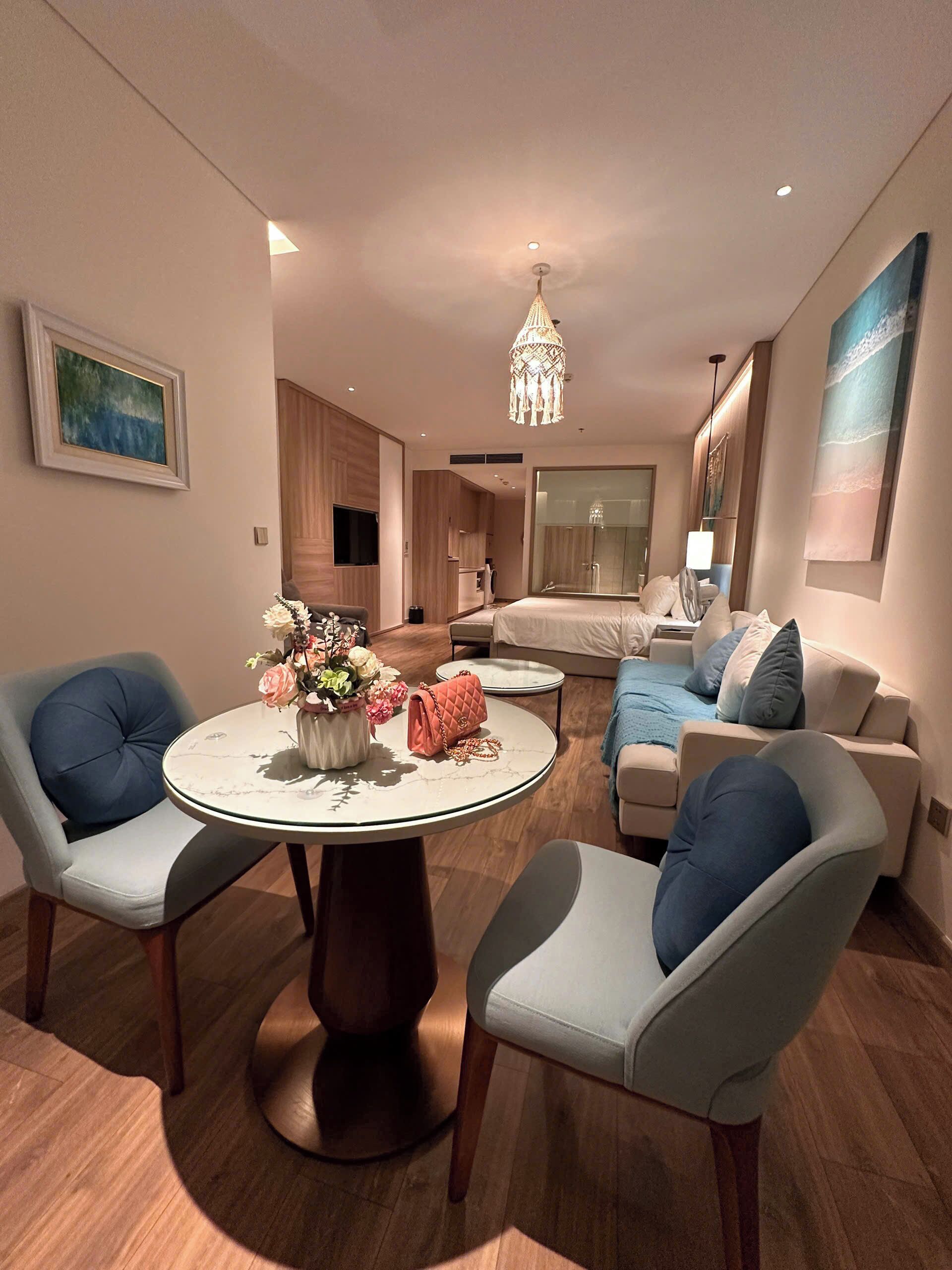 A modern hotel room with a round dining table, two armchairs, a sofa, a bed, a large window, and a chandelier.