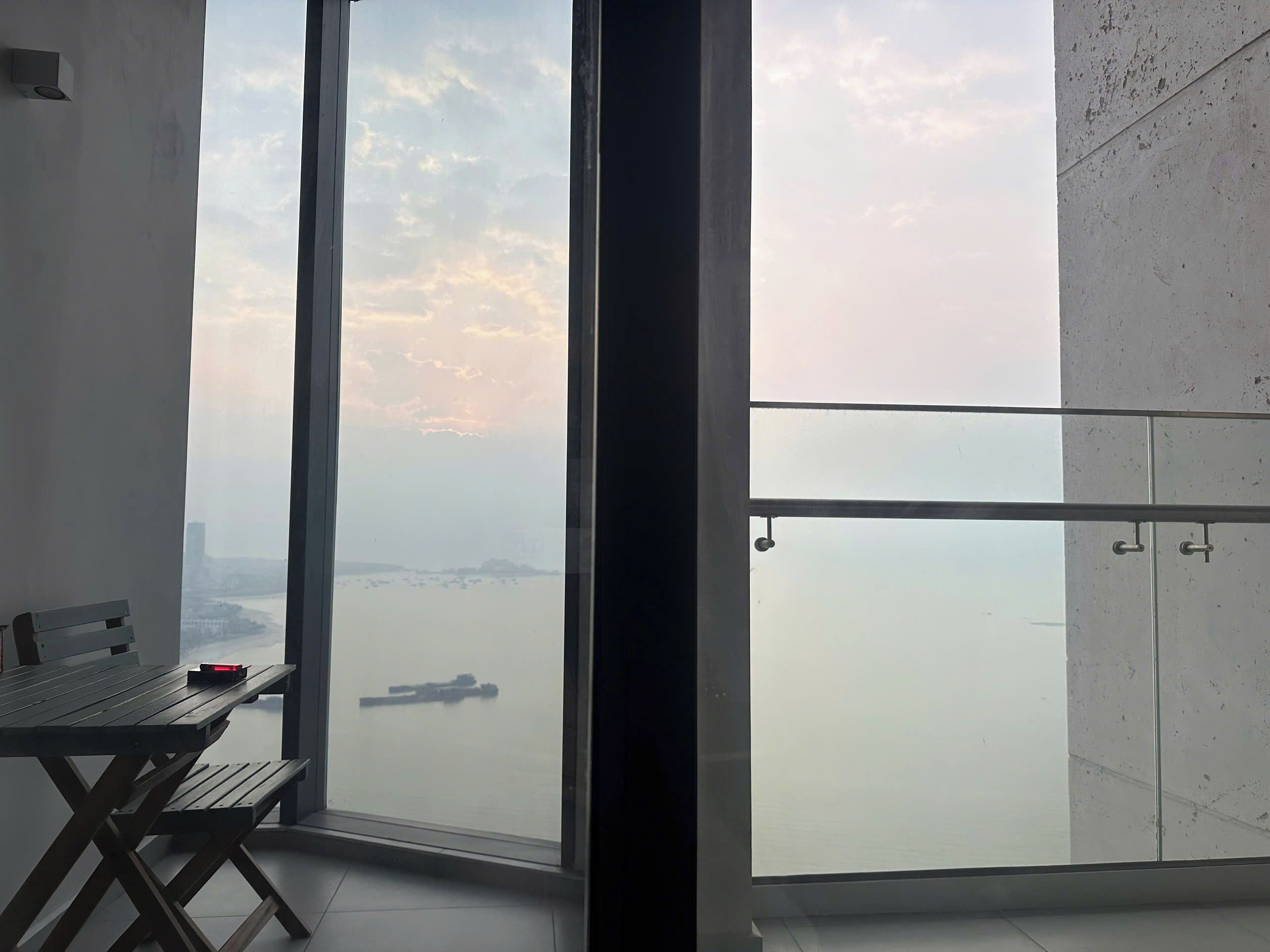 A balcony with a view of a body of water and distant land, a small table with a red object, and a bench.