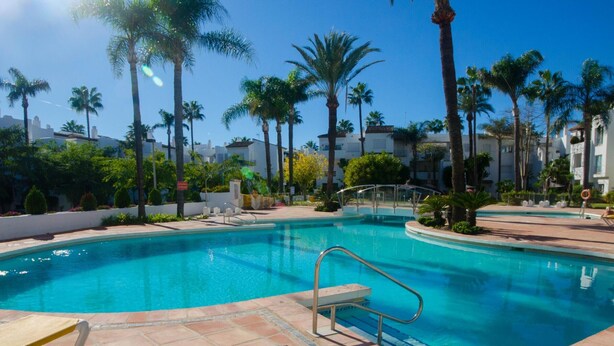 A swimming pool with a metal ladder, surrounded by palm trees and a tiled deck. - Spacious Apartment in Estepona with WiFi and Parking (Estepona)