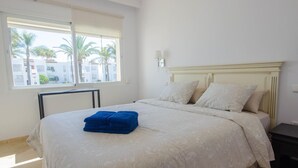 A hotel room with a bed, a nightstand, a window with a view of palm trees and buildings, and a wall-mounted lamp. - Spacious Apartment in Estepona with WiFi and Parking (Estepona)