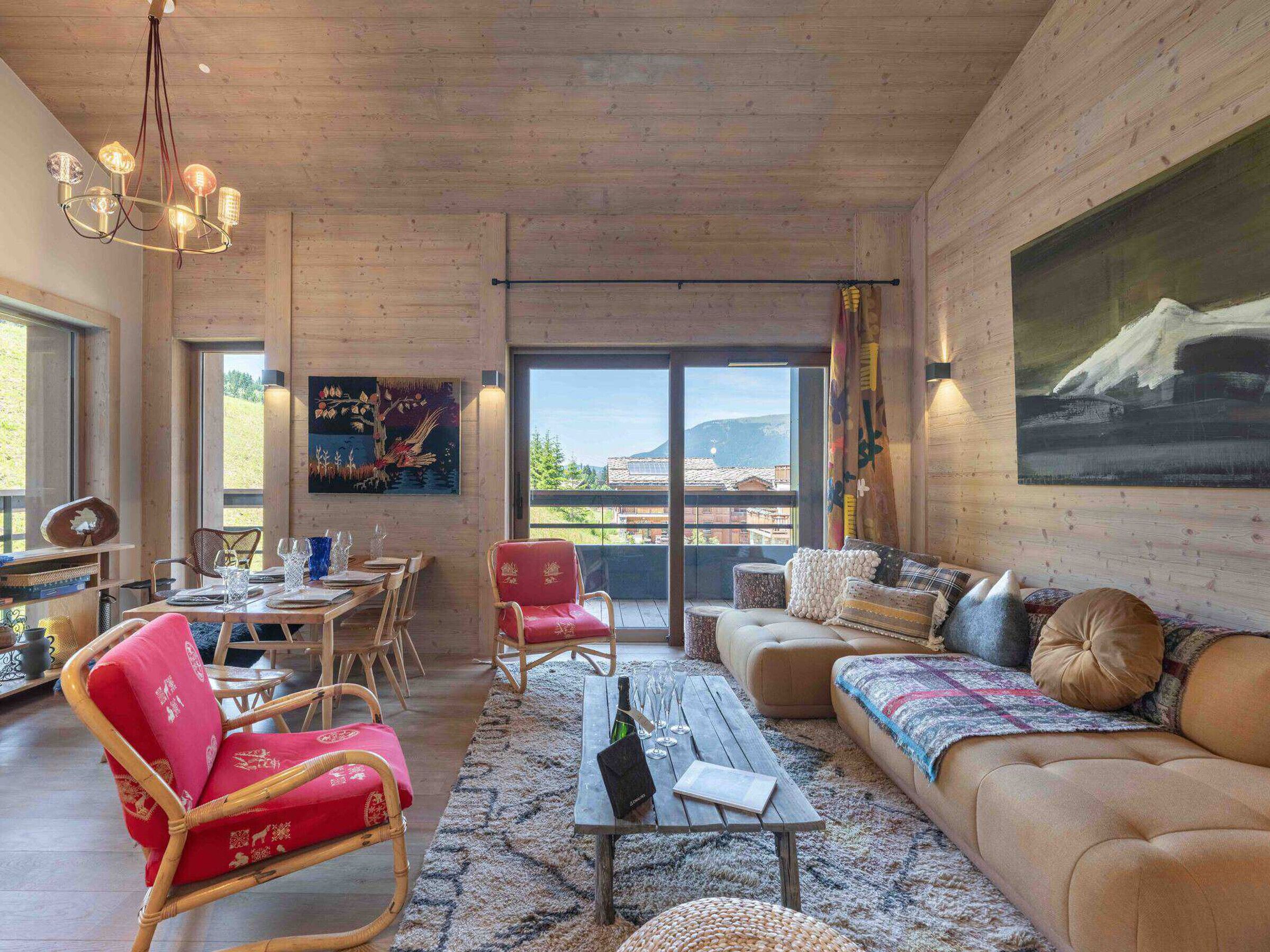 A spacious living area with a wooden ceiling, a dining table set for four, a red upholstered chair, a sofa with patterned cushions, and a large painting on the wall.