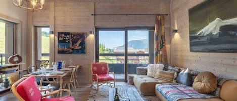 A spacious living area with a wooden ceiling, a dining table set for four, a red upholstered chair, a sofa with patterned cushions, and a large painting on the wall.
