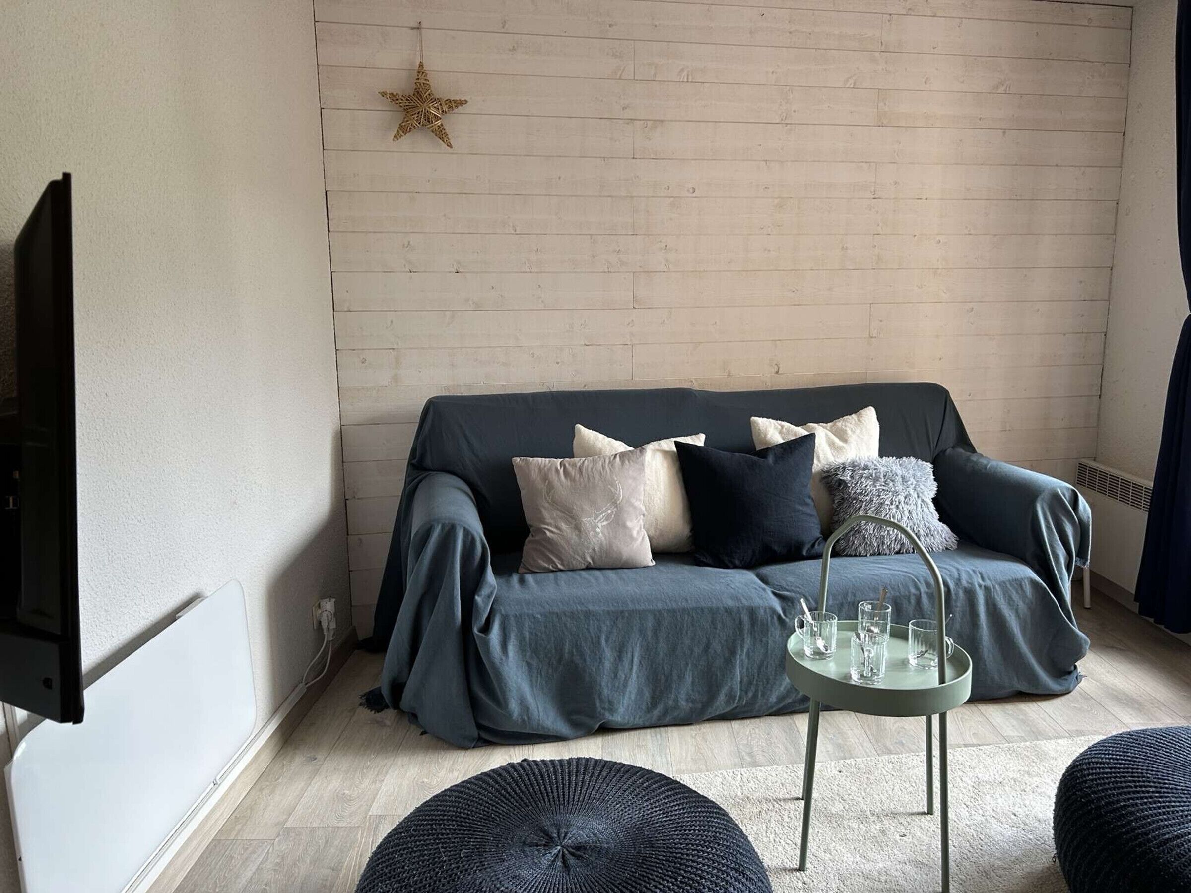 A modern living room with a dark blue sofa, multiple cushions, a small round table with glasses, and a star decoration on the wall.