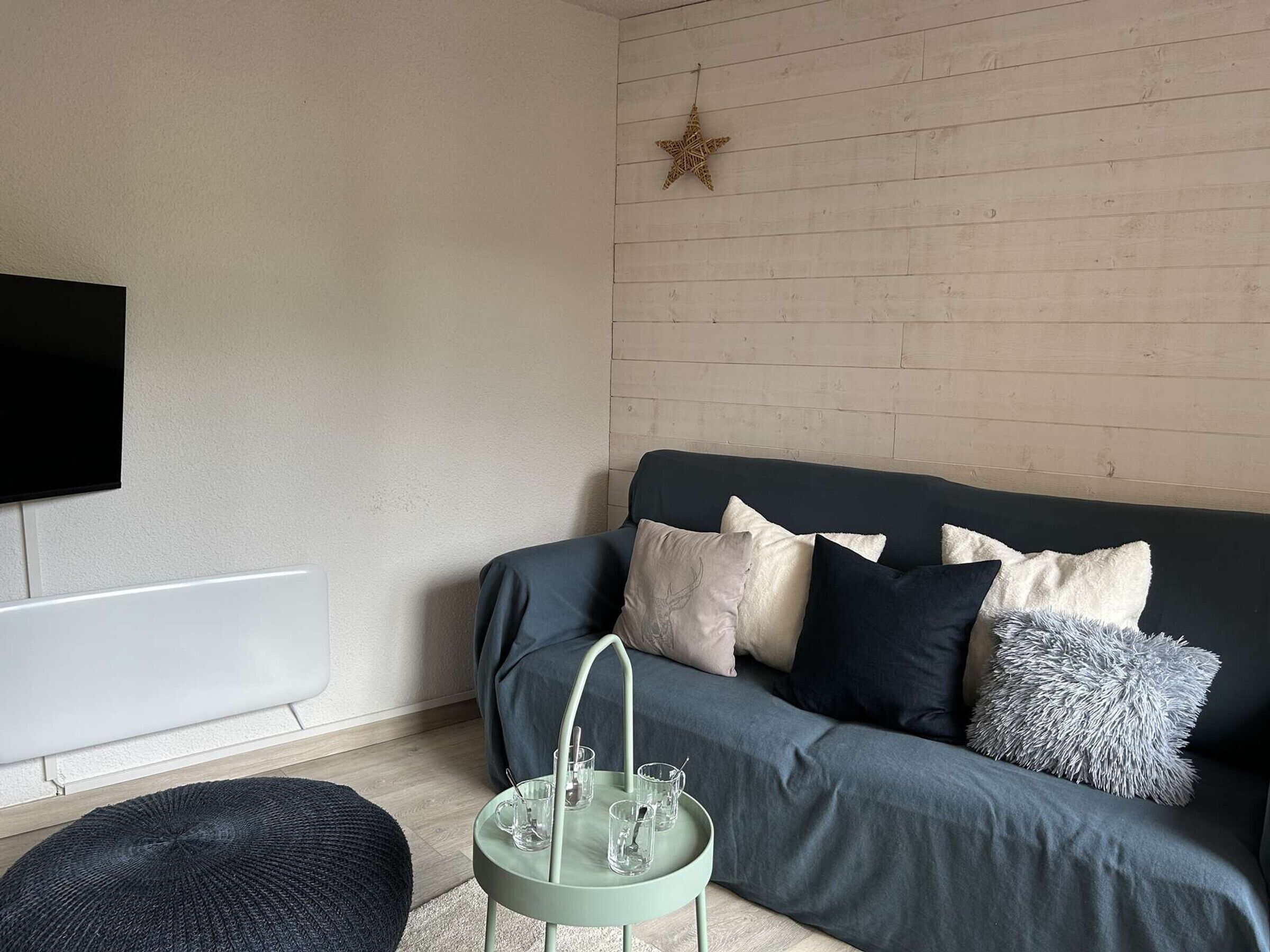 Un salon moderne avec un canapé gris, un pouf rond et une petite table d’appoint sur laquelle se trouvent des verres et une tasse.
