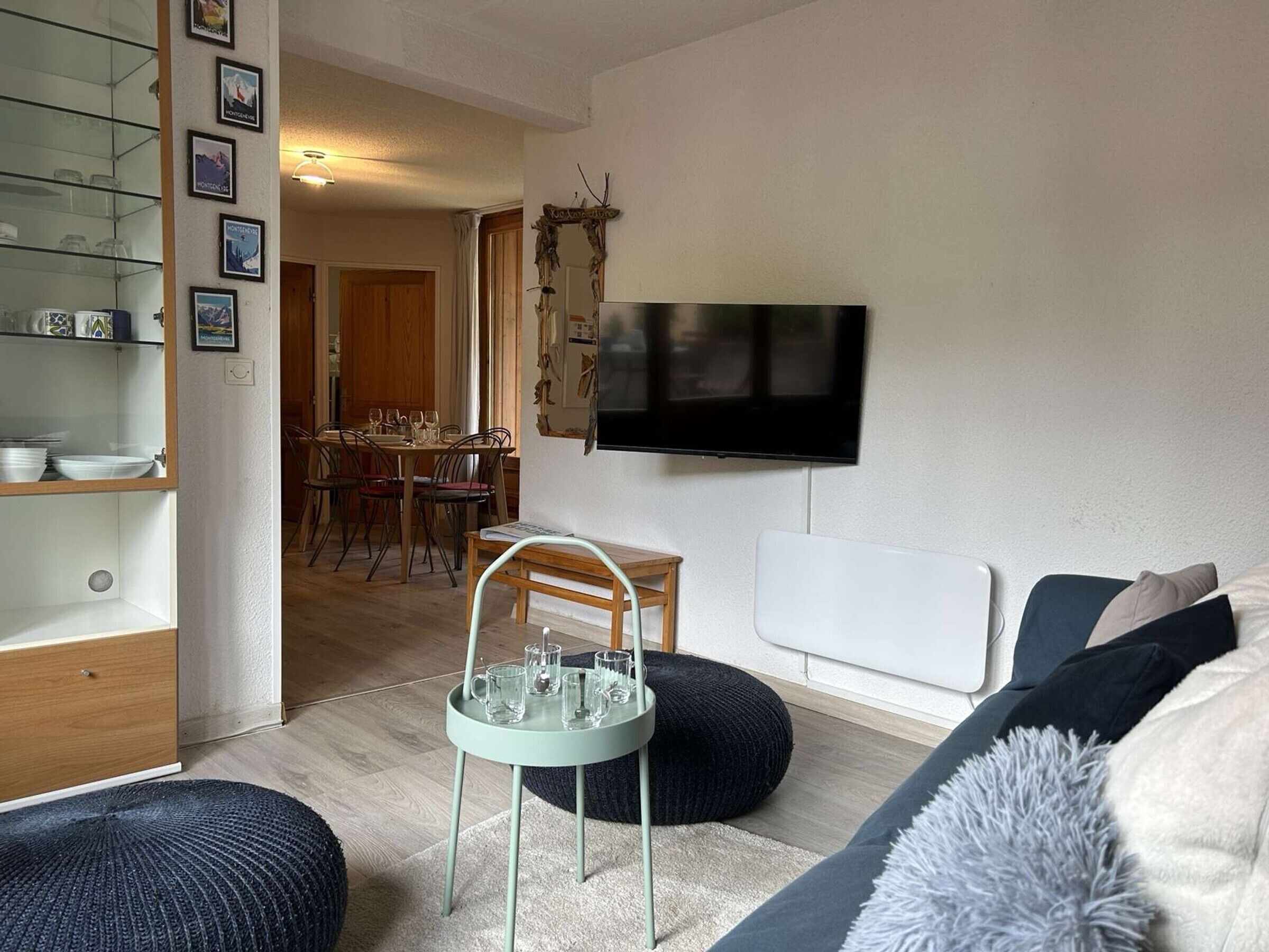 Ein Wohnzimmer mit an der Wand montiertem Fernseher, einem Essbereich mit Tisch und Stühlen sowie einer Sitzecke mit Sofa und Poufs.