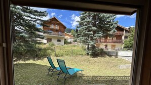 A view through a glass door of a building with a grassy yard, two blue chairs, and wooden buildings in the background. - Cozy Montgenèvre apartment, 4 pers, near center (Montgenèvre)