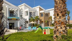 Un hôtel moderne à la façade blanche, doté d’un espace de jeux pour enfants avec un toboggan, et d’un palmier en premier plan.
