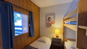 A small, cozy room with wooden walls, a single bed, a window with curtains, a bedside lamp, and a bunk bed.