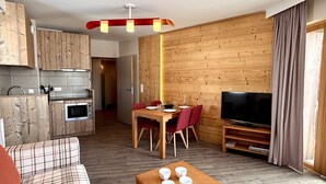 A modern living room with a wooden floor, a sofa, a dining table with red chairs, a TV, and a kitchen area with wooden cabinets and a microwave.