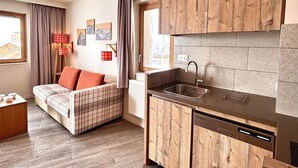 A compact living space with a kitchenette, a sofa with plaid bedding, and a view of the city through the window.