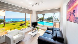 Un salón moderno con televisor, una mesa de centro blanca y un sofá de cuero negro. Amplias ventanas ofrecen vistas a una piscina y al mar.