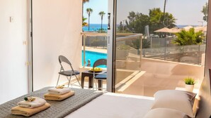 Un balcón con puerta de cristal que da a una zona de piscina, una cama con colchón acolchado gris, y una pequeña mesa con una silla.