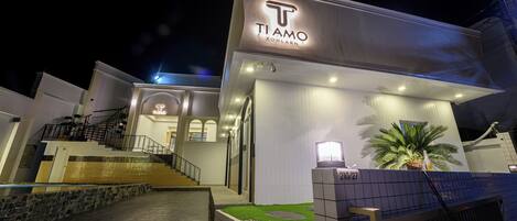 A modern hotel with a prominent 'TI AMO KOHLN' sign, a well-lit entrance, and a staircase leading to the upper floors.