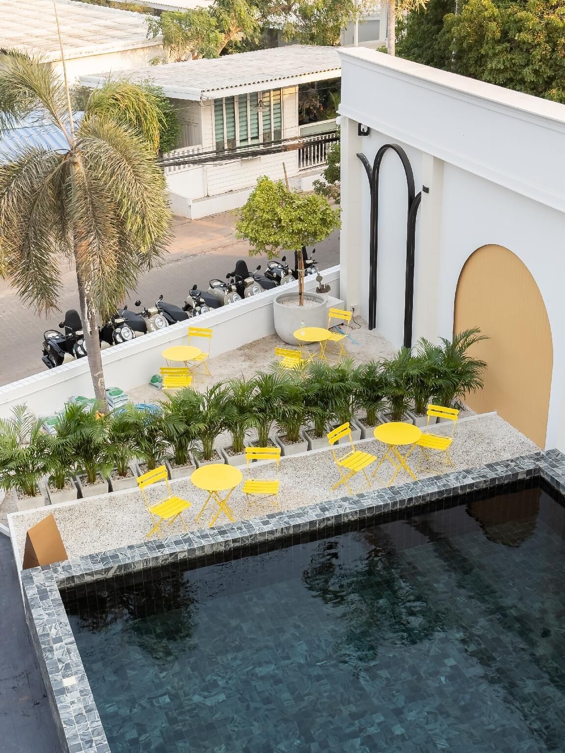 Un espace piscine aménagé sur le toit, avec des chaises et des tables jaunes, entouré de plantes en pot et offrant une vue sur les scooters garés.