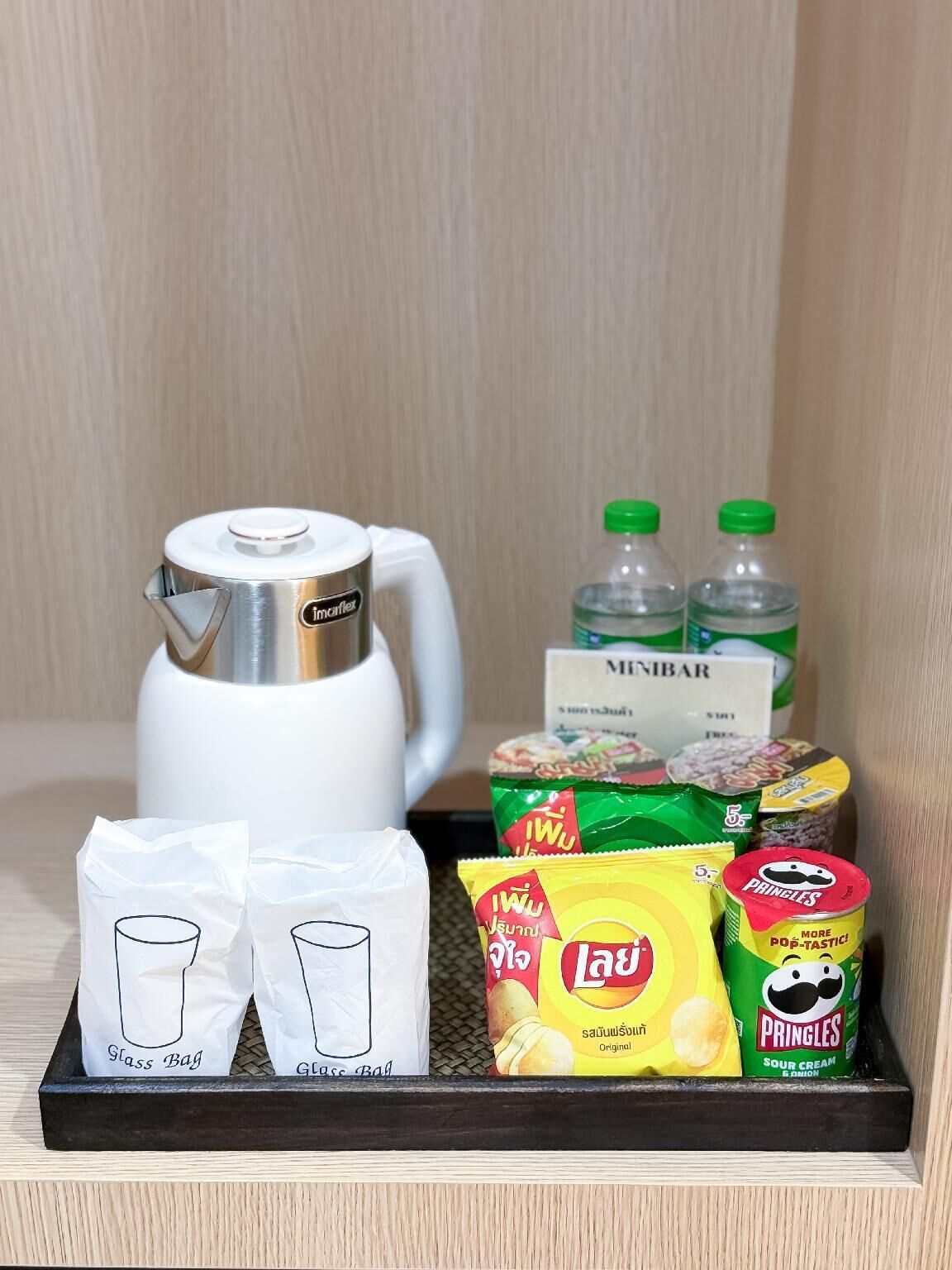 A tray with a white electric kettle, two bottles of water, a box of Minibar snacks, a packet of Lay chips, and two glass bags.