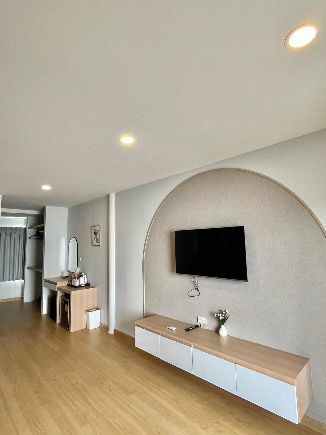 A modern hotel room with a wooden floor, a flat-screen TV mounted on a curved wall, a small desk with a mirror, and a bedside table with a vase of flowers.