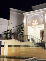 Entrada de un hotel con una escalera que conduce al edificio principal, un letrero de neón sobre la entrada y un pequeño área de piscina al frente.