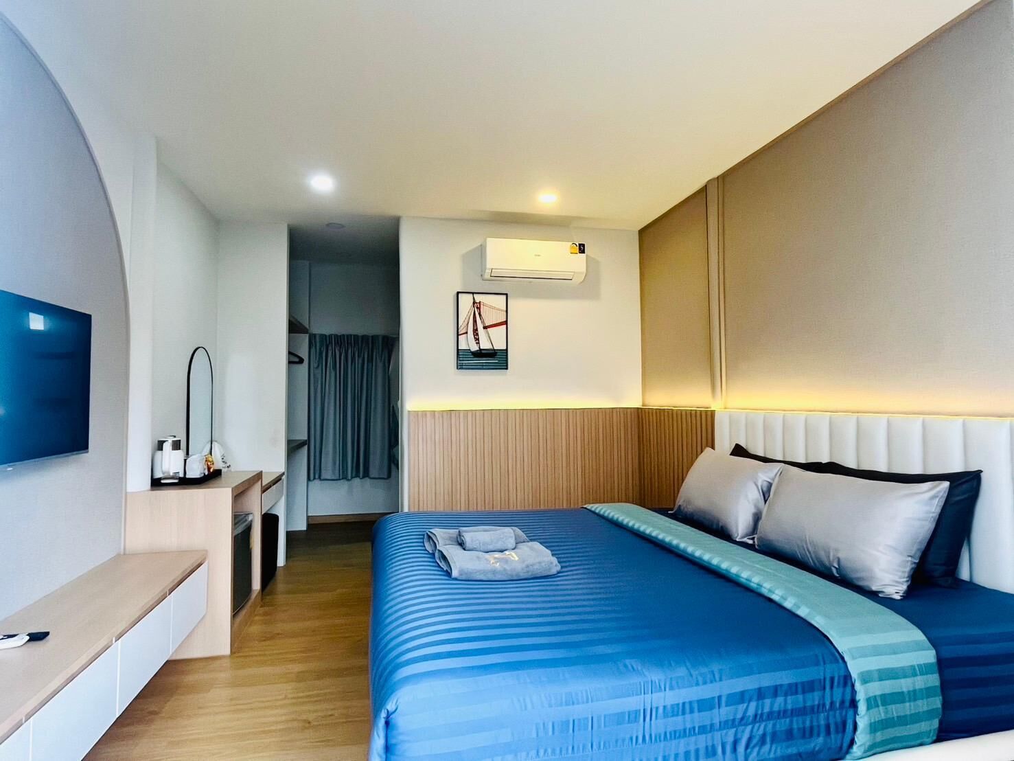 A modern hotel room with a large bed, a flat-screen TV, a desk with a mirror, and an air conditioning unit.