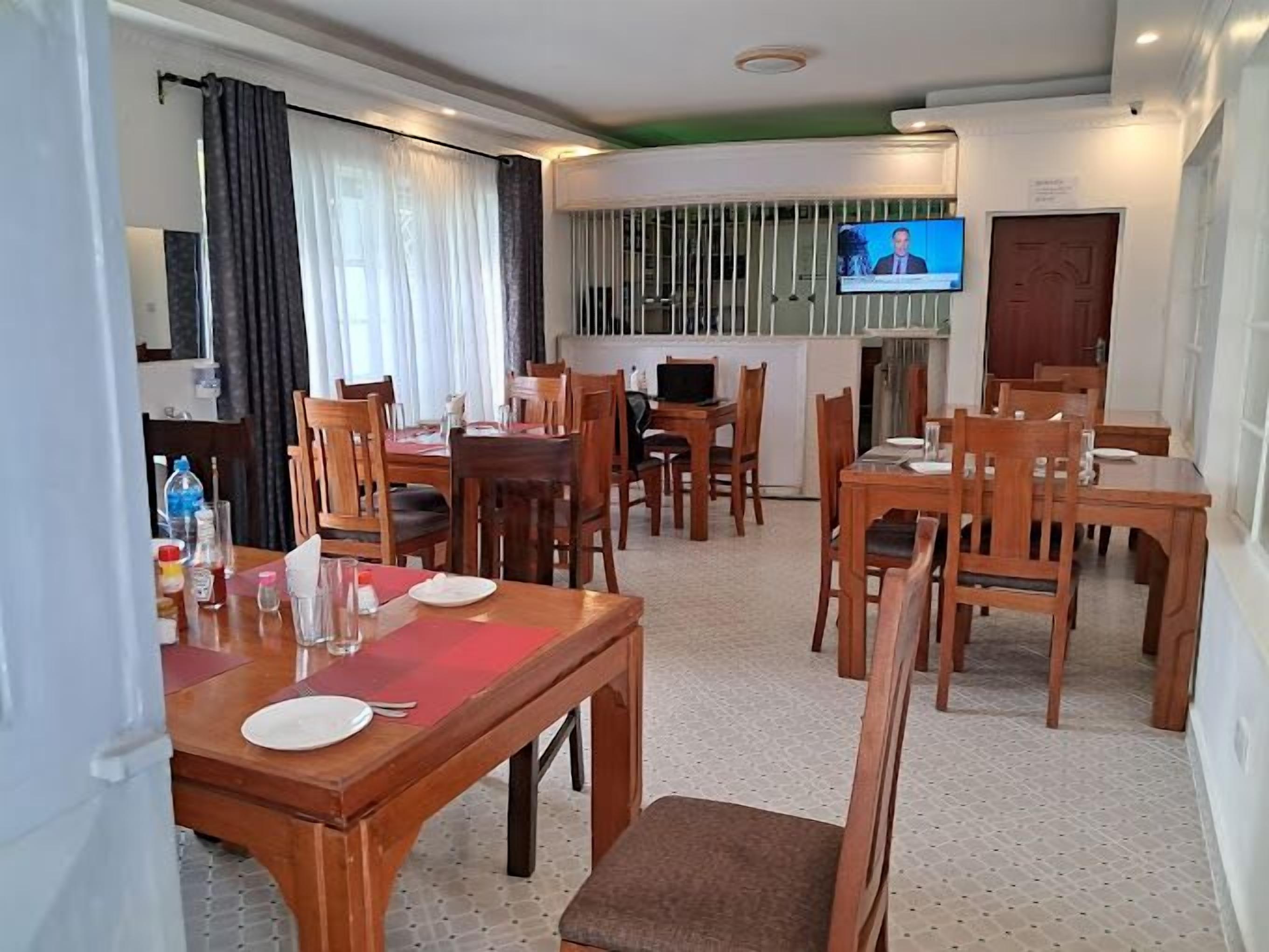 Une salle à manger d’hôtel avec des tables et des chaises en bois, un téléviseur fixé au mur et une fenêtre avec des rideaux.