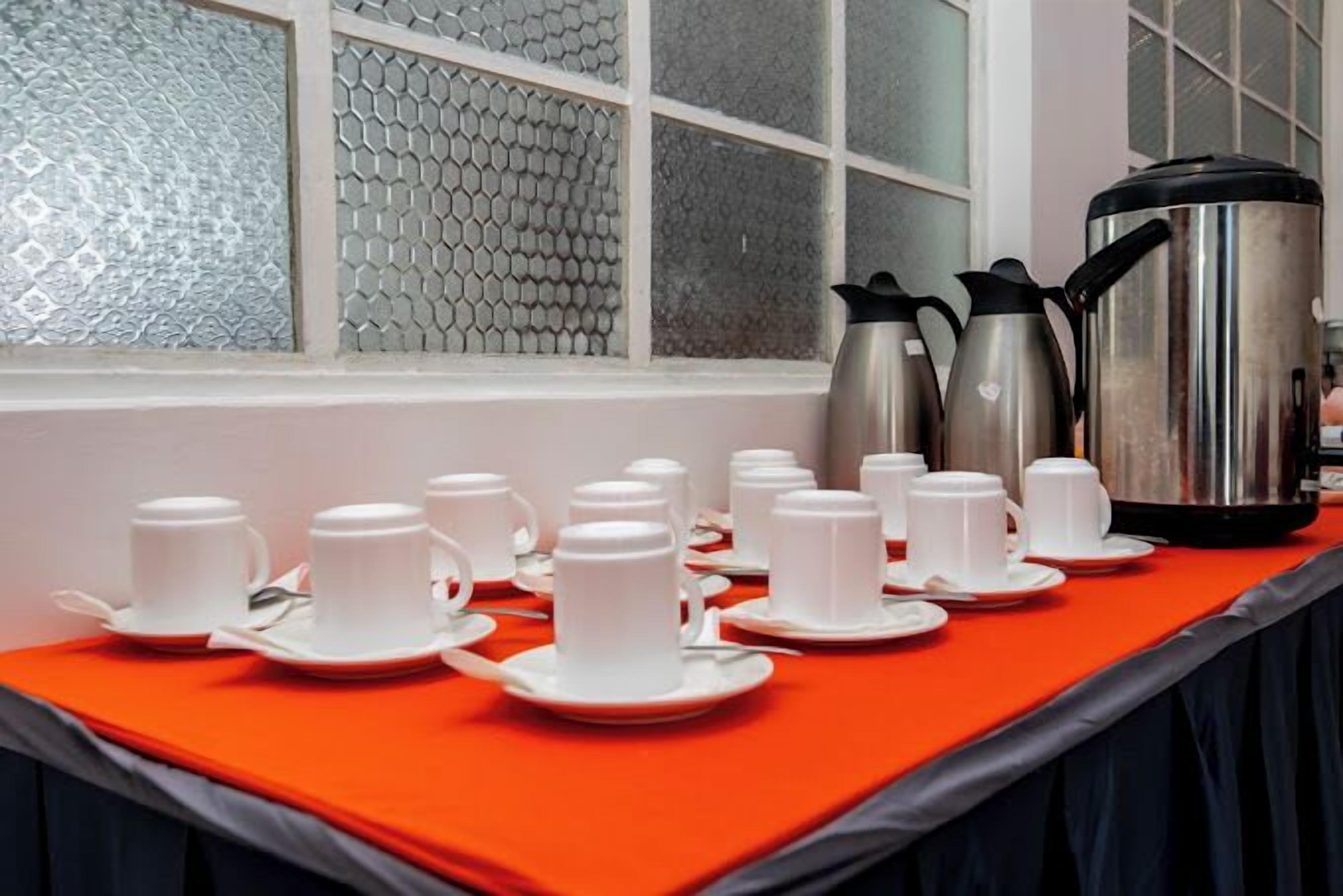 A breakfast setup with multiple white cups and saucers on a red tablecloth, a silver coffee pot, and a smaller electric kettle.