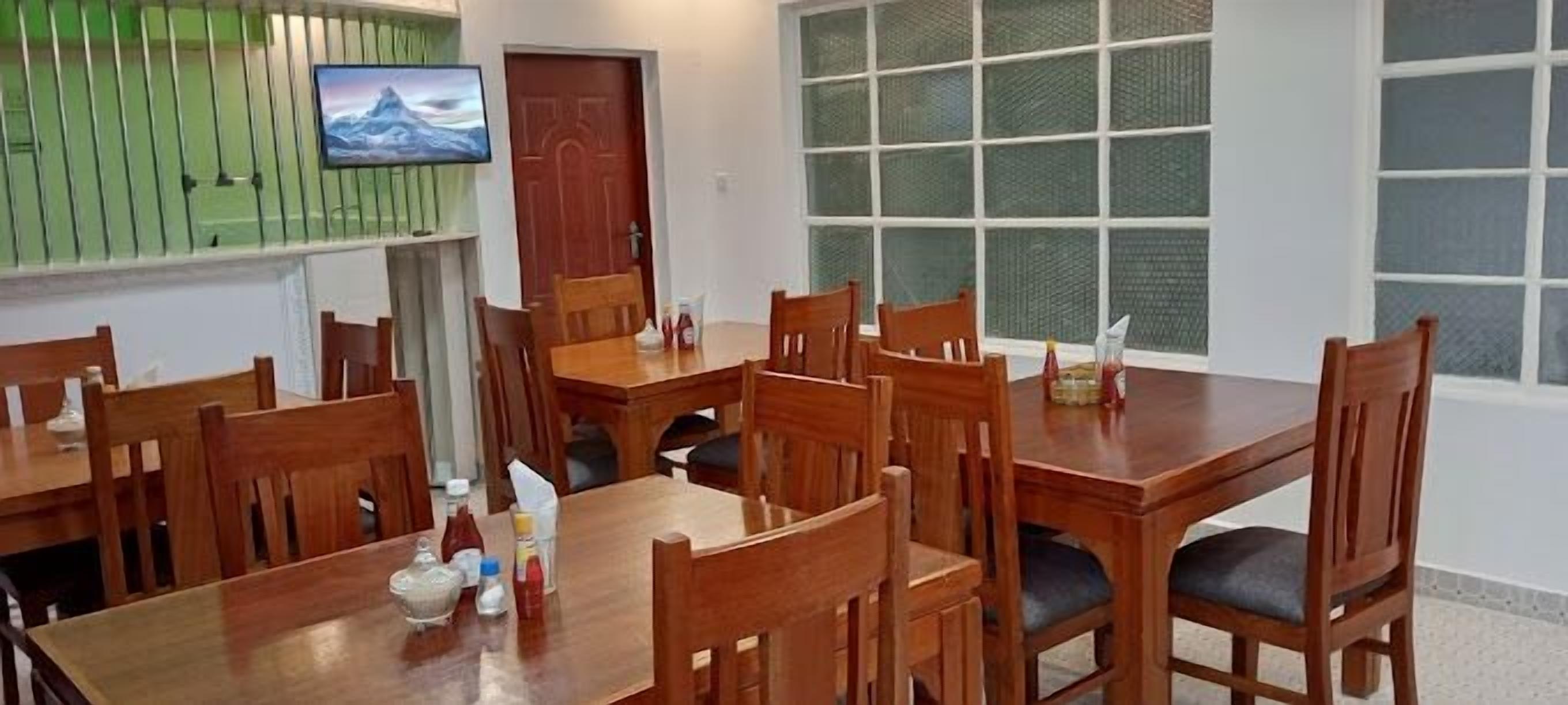 Un comedor con mesas y sillas de madera, un televisor montado en la pared y un ventanal con vidrios satinados.