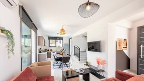 Un salon moderne avec un canap é, une table basse et un coin repas avec des chaises. On aperçoit au fond une cuisine, un escalier et un téléviseur fixé au mur.