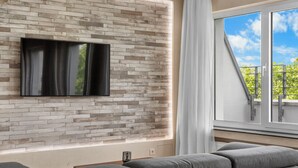A modern living room with a brick-patterned wall, a flat-screen TV, a grey sofa, and a large window with a view of greenery. - Domloge Lofts & Apartments (Cologne)