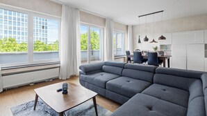 A modern living room with a sectional sofa, a wooden coffee table, and a view of the outdoors through large windows. - Domloge Lofts & Apartments (Cologne)